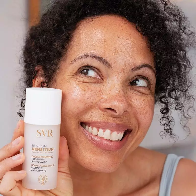 Femme aux cheveux bouclés souriante tenant le flacon blanc SVR Densitium Bi-Sérum contre sa joue par