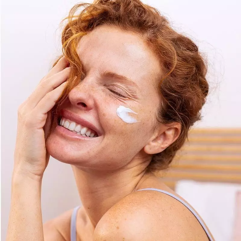 Femme rousse souriante aux taches de rousseur appliquant la Crème Densitium Riche SVR sur sa joue, t
