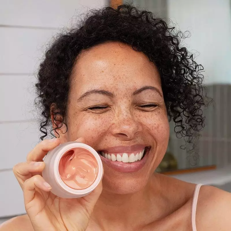 Femme souriante aux taches de rousseur tenant le pot SVR DENSITIUM Rose Éclat ouvert, révélant la te
