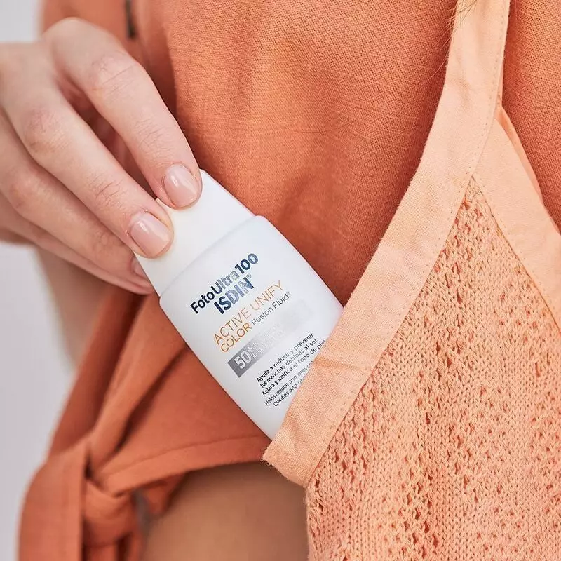 Femme tenant le tube ISDIN Fotoultra 100 Active Unify Color SPF 50+ dans la poche de son pull orange