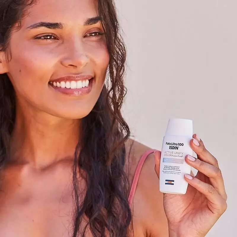 Femme souriante tenant le tube ISDIN Fotoultra 100 Active Unify Color SPF 50+ près du visage, en cad