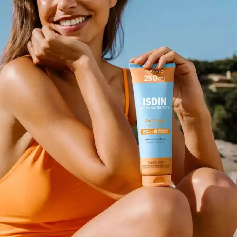 Femme souriante en maillot de bain orange tenant le tube ISDIN Gel Crème SPF 50+ 250ml sur plage ens