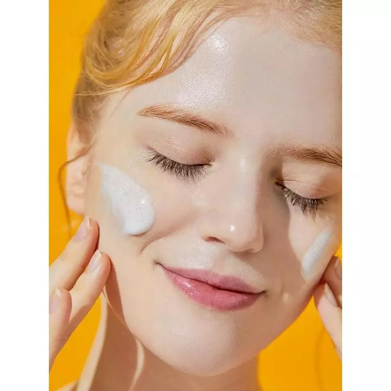 Femme blonde souriante avec des mousses de Nettoyant Gel COSRX à la Mucine d'Escargot sur les joues,