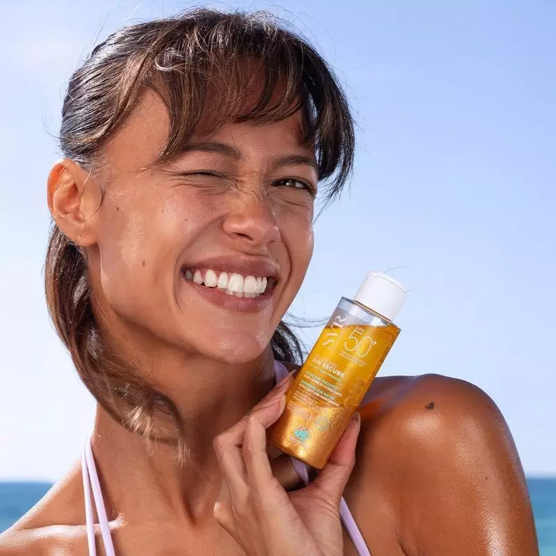Femme souriante à la plage tenant le flacon SVR Sun Secure Glow SPF50+, peau bronzée glow et effet s