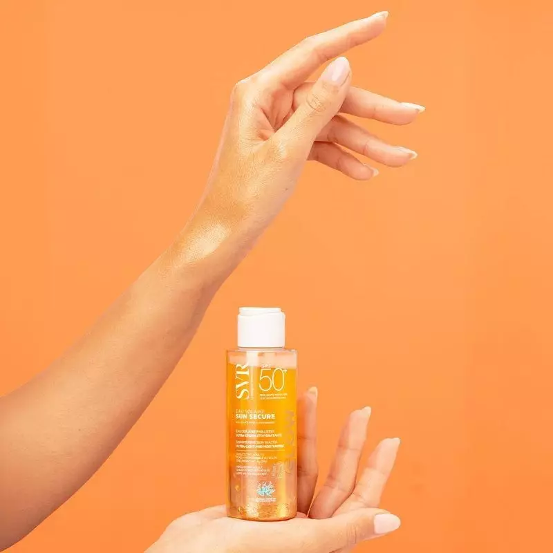 Main tenant le flacon SVR Sun Secure Glow SPF50+ sur fond orange, liquide doré scintillant et peau d