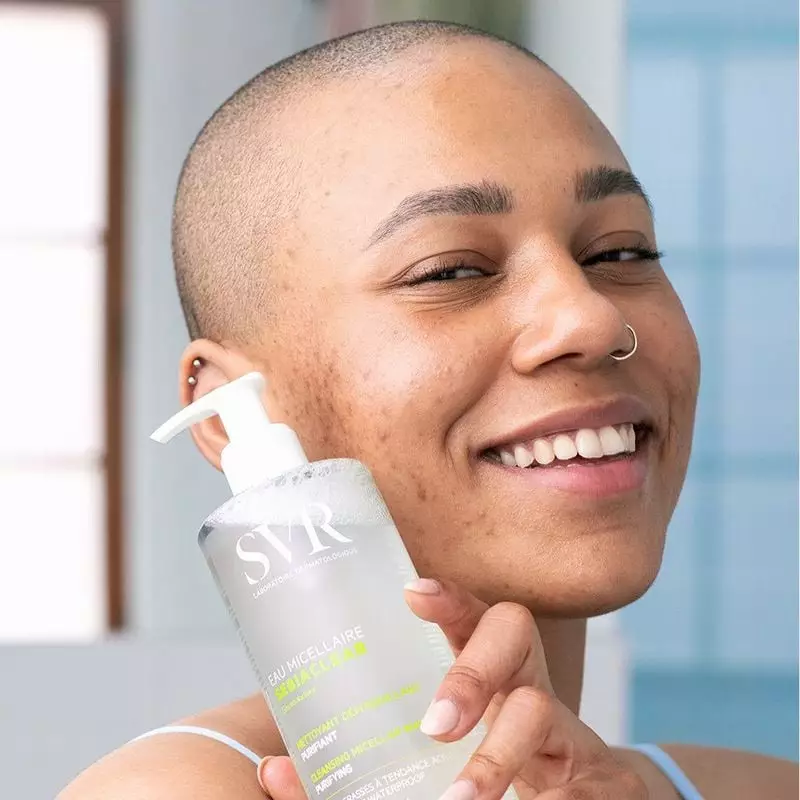 Modèle aux cheveux courts souriante en salle de bain tenant la bouteille d'eau micellaire SVR Sebiac