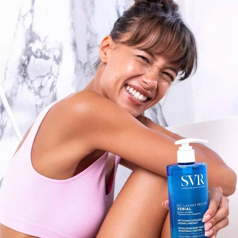 Femme souriante tenant un flacon de SVR Xérial Gel Lavant Peeling dans une salle de bain.