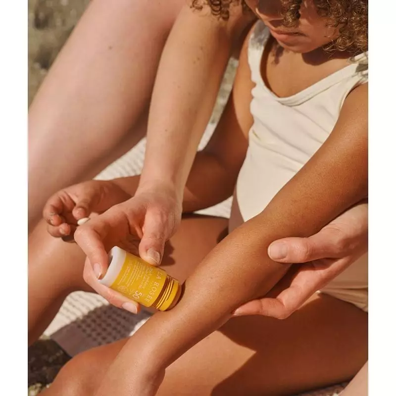 Mère appliquant le Stick Solaire SPF 50+ La Rosée sur le bras de l'enfant sur serviette de plage en 