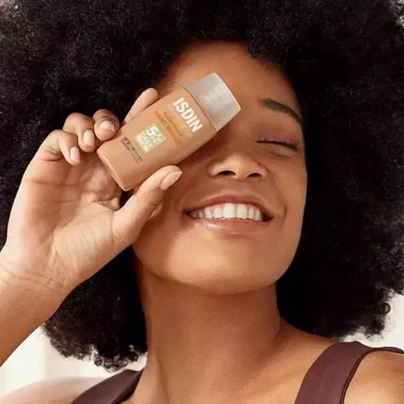 Femme souriante aux cheveux bouclés appliquant l'ISDIN Water Color Bronz SPF50 teinté bronze près de