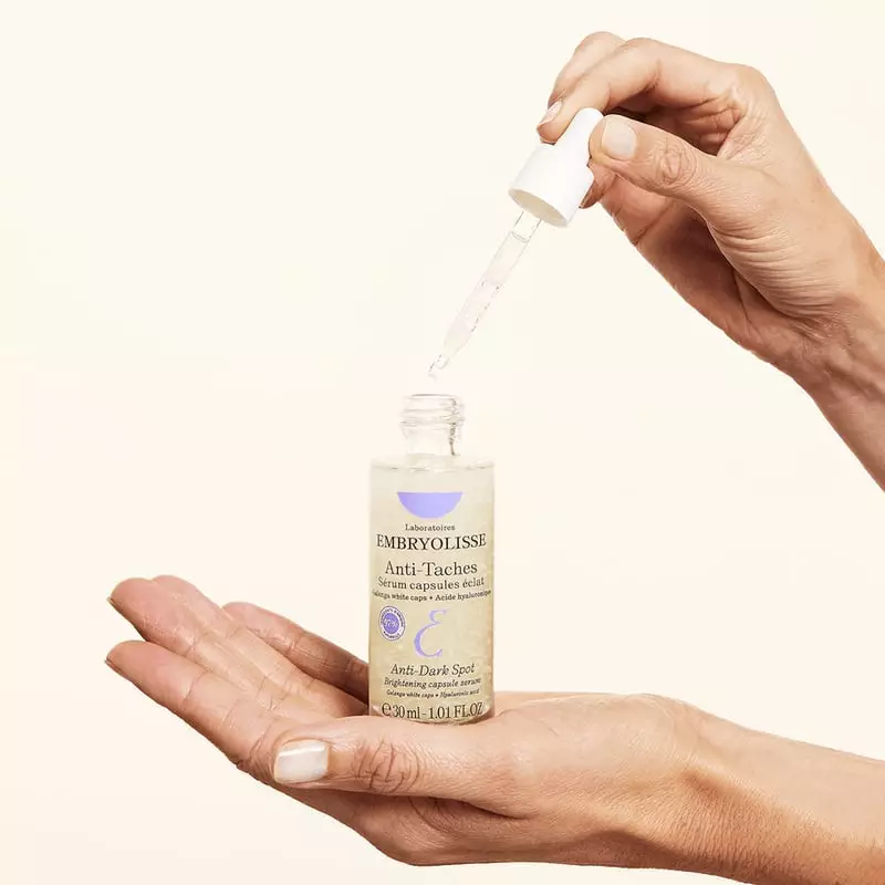Femme tenant le flacon de sérum Embryolisse près de son visage souriant, démontrant le produit en ca