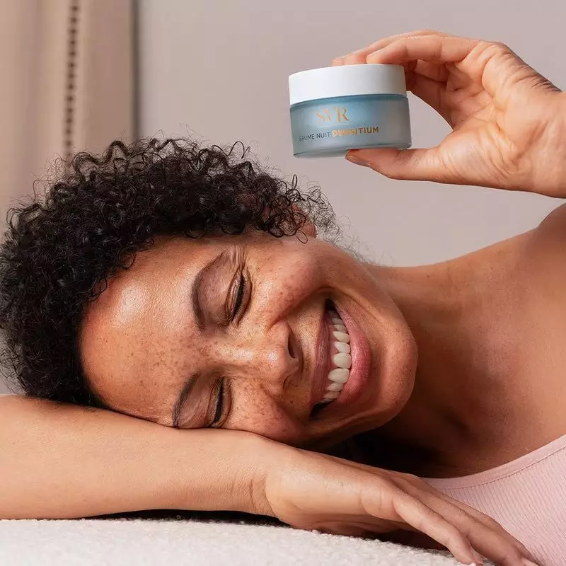 Une femme tient le pot SVR Densitium Baume Nuit Recharge, en souriant.