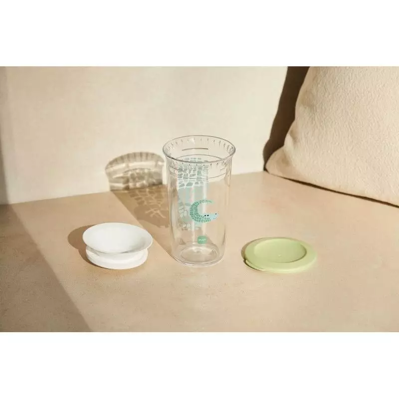 Petite fille tenant un verre d'apprentissage MAM 360° vert et buvant, souriant dans le salon.