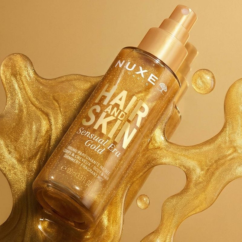 Femme vaporisant la brume corps et cheveux NUXE Sensual Era Gold, créant une aura scintillante.