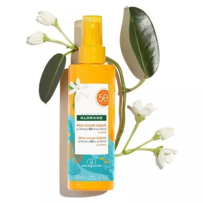 Flacon de spray solaire Klorane SPF50 avec fleurs de tiaré blanches et feuilles vertes sur fond beig