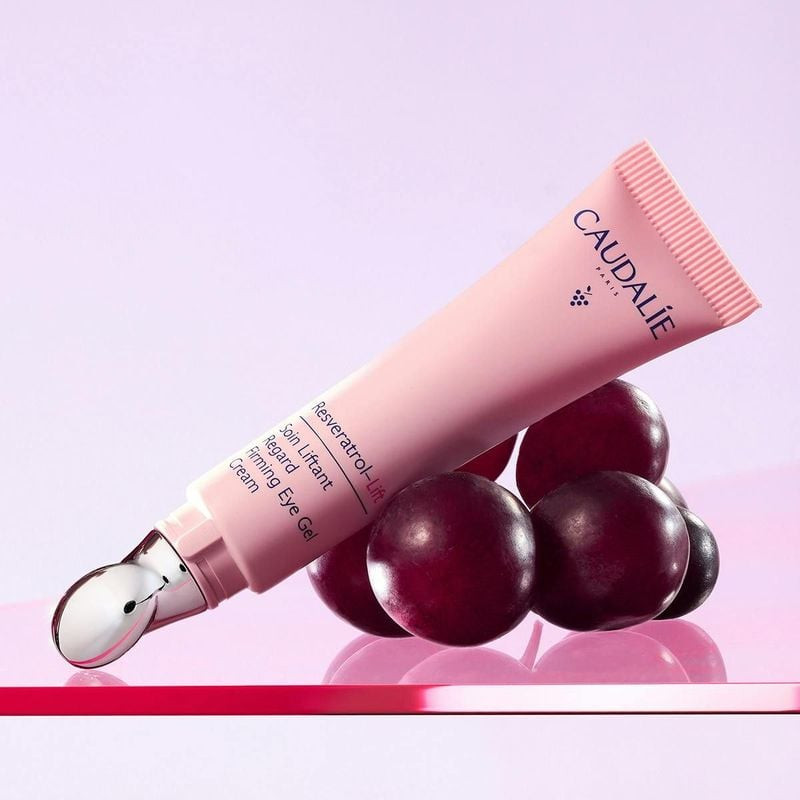 Tube de soin liftant regard Caudalie Resvératrol Lift posé sur des raisins rouges foncés, fond rose 