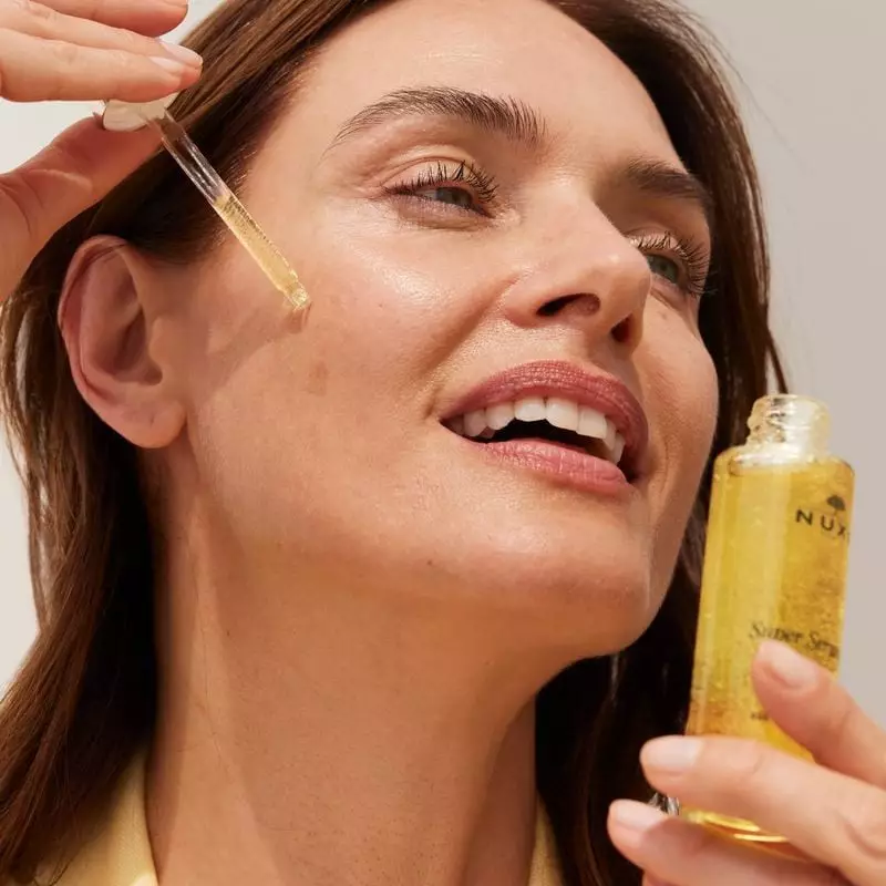Femme souriante appliquant Nuxe Super Sérum [10] Longevity sur sa joue avec une pipette