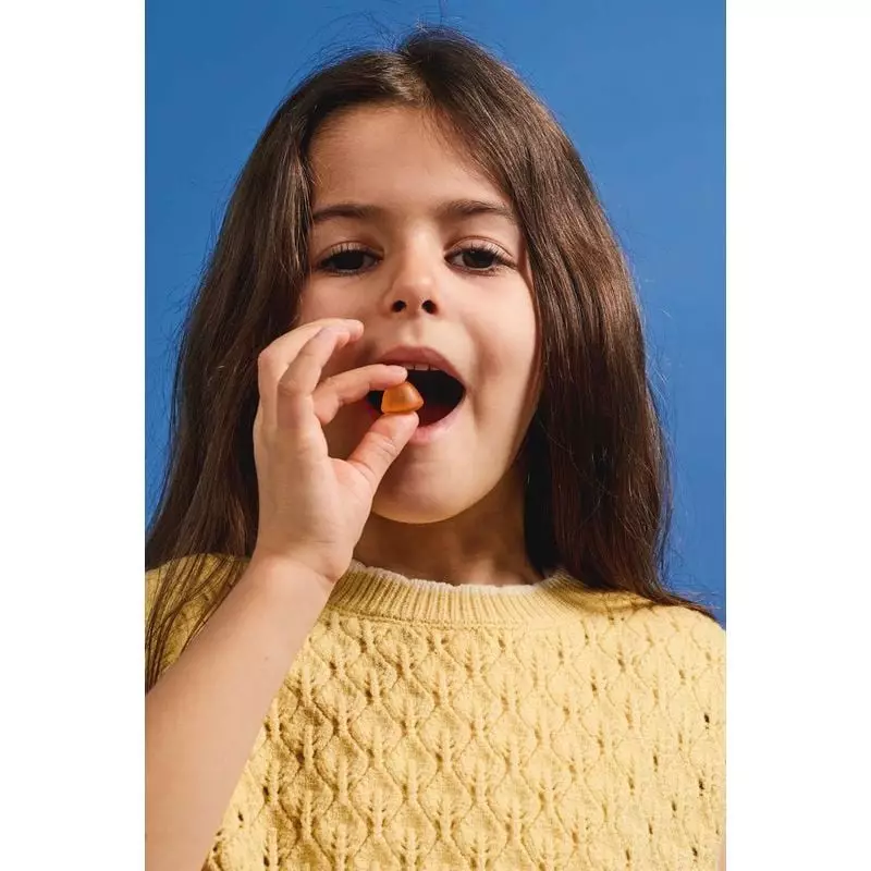 Jeune fille mangeant une gomme orange Mium Lab Safran B6 pour enfants sur fond bleu.